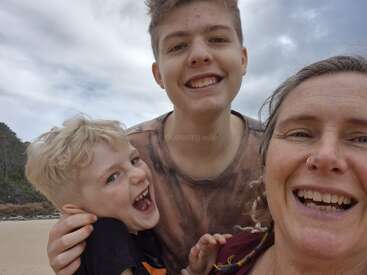 A joyful selfie of a woman and two boys at the beach. They’re smiling and laughing, enjoying a fun moment together under a cloudy sky.