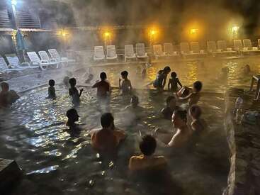 Por la noche, la gente se relaja en una piscina termal iluminada y llena de vapor. Las tumbonas se alinean en la terraza, mientras los grupos charlan y disfrutan juntos del ambiente cálido y brumoso.