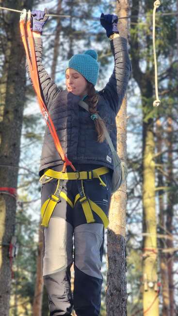 Una mujer vestida con ropa de montaña y un sombrero turquesa está escalando o lanzándose en tirolina en un parque de aventuras en el bosque, sujeta por arneses de seguridad y cuerdas.