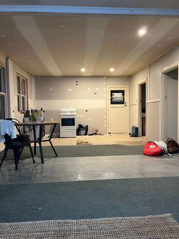 Una habitación parcialmente renovada, con paredes de yeso sin terminar, iluminación básica, una cocina blanca, una mesa con sillas, bolsas esparcidas por el suelo y un mobiliario mínimo, que muestra signos de obras en curso.