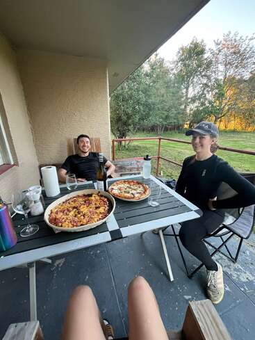 Tres amigos están sentados juntos en una terraza, disfrutando de una pizza casera y unas bebidas en una mesa. El ambiente es relajado, rodeados de vegetación y la luz del atardecer.