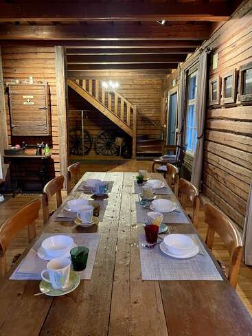 Ein rustikales Esszimmer aus Holz mit einem langen Tisch für acht Personen, mit bunten Tassen, Schüsseln, Tischsets, Holzstühlen, Holzwänden und einer Treppe im Hintergrund.