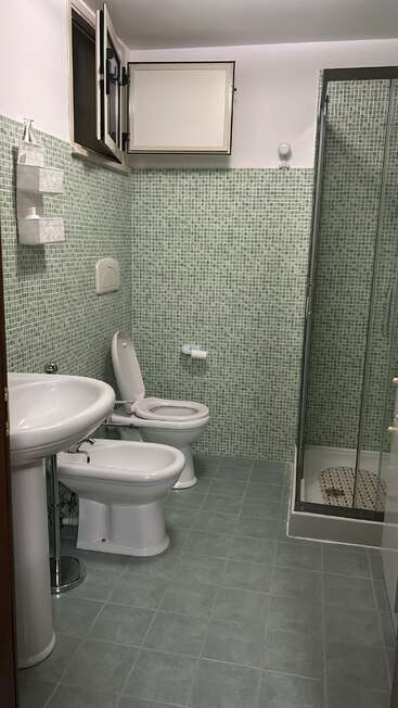 Das ist ein gepflegtes Badezimmer mit grünen Mosaikfliesen, ausgestattet mit Waschbecken, Toilette, Bidet, einer Dusche mit Glastüren, einem Fenster, das sich öffnen lässt, und an der Wand befestigten Ablagekörben.