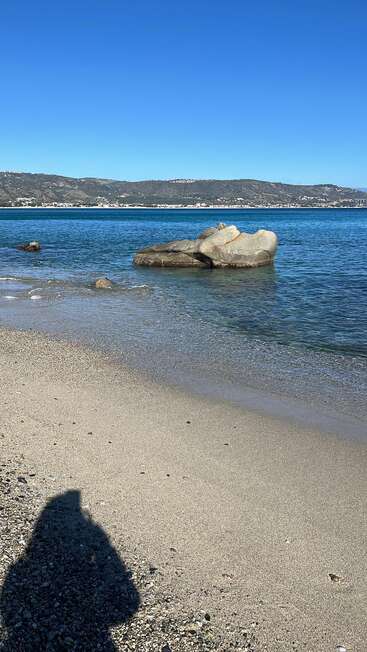 Ein Sandstrand mit klarem, blauem Wasser, großen, teilweise im Wasser liegenden Felsen, fernen Hügeln und einer Stadt sowie dem Schatten einer Person im Vordergrund.