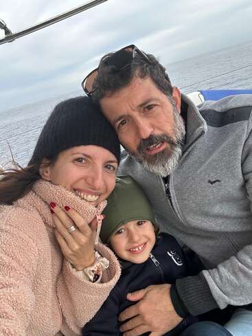 Eine lächelnde dreiköpfige Familie genießt einen gemütlichen Moment im Freien, warm eingepackt auf einem Boot, mit dem Meer und dem bewölkten Himmel im Hintergrund.