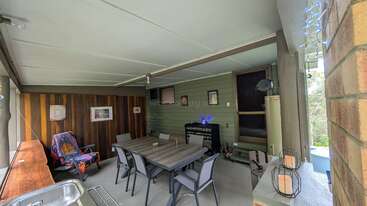 This image shows a cozy covered patio with a wooden dining table, six chairs, armchair, wall art, string lights, brick accents, and various decorations.