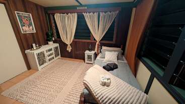 This cozy bedroom features a single bed, neutral-colored decor, a textured rug, wooden walls, soft lighting, potted plant, artwork, sheer curtains, and decorative towels.