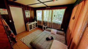 Este acogedor dormitorio presenta paredes con paneles de madera, una cama baja con mantas, grandes ventanas con cortinas transparentes, una pequeña cómoda blanca, plantas, iluminación cálida y toques decorativos.