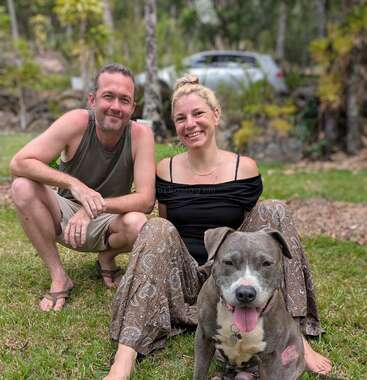 A man and woman are sitting on the grass outside, smiling happily. A gray and white dog sits in front of them, also looking content.