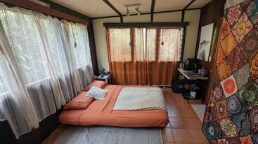 This cozy bedroom features large windows with sheer curtains, an orange bed, a colorful quilt, tiled floor, small desk, coffee station, and natural light throughout.