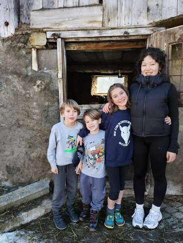 Four smiling children stand together in front of an old, rustic wooden building. They wear casual clothes and look happy, enjoying a day outdoors together.
