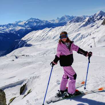 A woman in a pink ski suit smiles on a snowy mountain, holding ski poles. Majestic snow-covered peaks and a clear blue sky surround her.