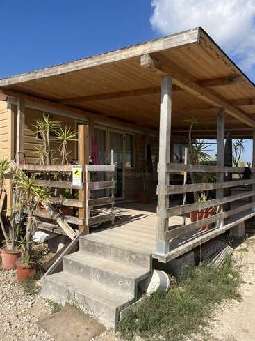 The image depicts a wooden house with a covered porch and ramp, featuring potted plants and a gravel driveway, set against a blue sky with white clouds.