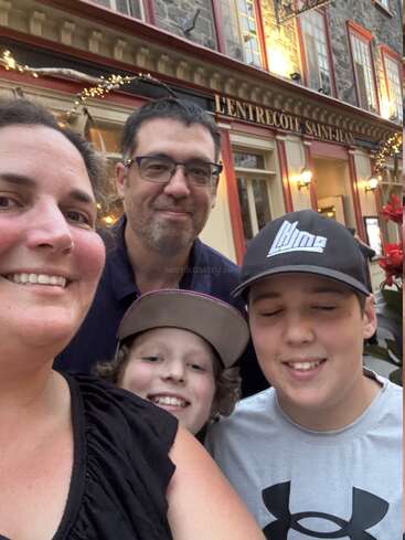 A smiling family of four poses for a selfie outside L’Entrecote Saint-Jean restaurant. Festive lights and a charming building create a warm, joyful atmosphere.