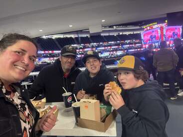 A family of four enjoys food and drinks at a stadium event. Smiling for a selfie, they sit near a counter with sports screens in the background.