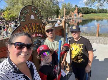 A smiling family of four poses in front of a mini golf sign by a pond. They appear happy, holding putters, enjoying a sunny outdoor day together.