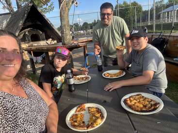 A family of four enjoys pizza and soda at an outdoor table on a sunny day. Everyone looks happy, sharing a meal by a woodfire oven.