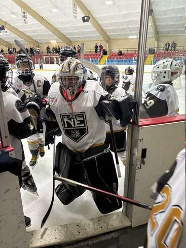 A youth hockey team, dressed in white and black uniforms, gathers at the rink entrance. The goalie leads, holding the door, ready for action.