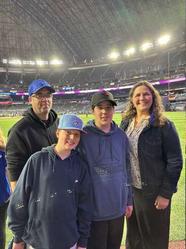 Una familia de cuatro miembros posa junta en un campo de béisbol, sonriendo. Las luces del estadio brillan intensamente sobre ellos y, al fondo, se ven las gradas.