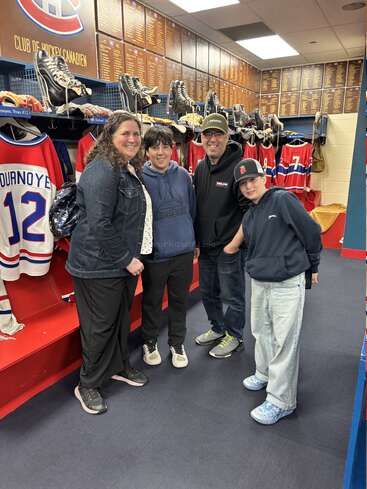 Una familia de cuatro miembros posa y sonríe junta en el vestuario de un equipo de hockey, repleto de camisetas, patines y placas conmemorativas, mientras disfrutan de una visita a unas instalaciones deportivas.