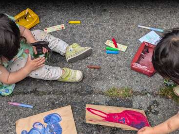 Duas crianças estão sentadas na calçada, rodeadas de materiais de arte, pintando desenhos coloridos em sacos de papel pardo. Giz de cera, canetas hidrográficas e um borrifador estão espalhados ao redor delas.