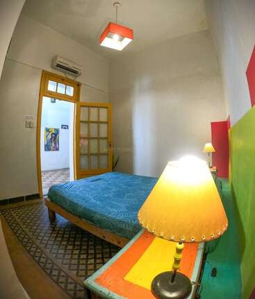 Un acogedor dormitorio con cama azul, cabecero de colores, lámparas de mesilla, suelo estampado, puerta amarilla, paredes blancas y un cuadro que se ve a través de la puerta abierta.