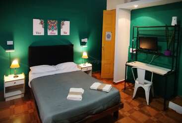 Un acogedor dormitorio con una pared de acento verde, cama de matrimonio, mesillas de noche, lámparas, pequeño escritorio con silla, TV, planta y arte mural "Vive Ama Rie".