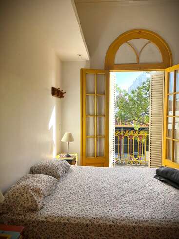 Un dormitorio luminoso con ropa de cama de flores, una lámpara de mesilla de noche, puertas francesas de marco amarillo que se abren a un balcón, dejando entrar la luz del sol, y verdes árboles que se ven en el exterior.