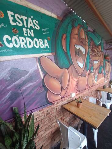La imagen muestra un vibrante mural en una pared con caras risueñas de dibujos animados, un cartel en el que se lee "ESTÁS EN CÓRDOBA" y sencillas mesas de madera con sillas blancas.