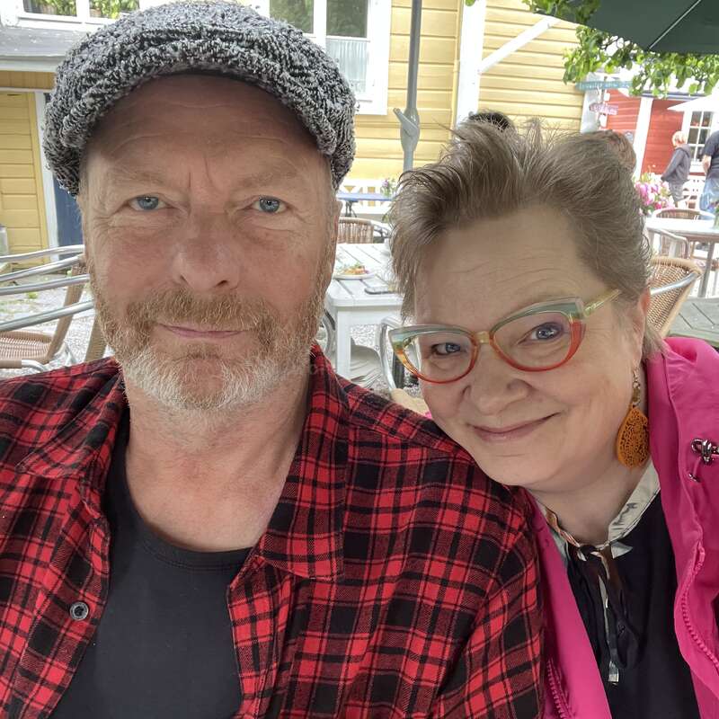 Une femme souriante aux lunettes colorées et un homme portant une casquette plate et une chemise à carreaux rouges posent ensemble en plein air, semblant heureux et détendus dans un café.