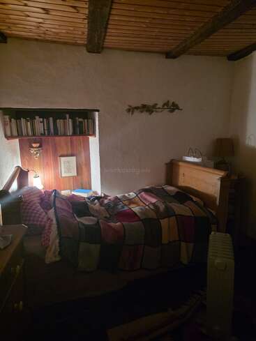 Ein gemütliches, schummriges Schlafzimmer mit einer Patchworkdecke auf dem Bett. Holzdecke, Bücherregal, Wanddekoration, Lampe, Kommode und ein tragbarer Heizkörper sorgen für Wärme.