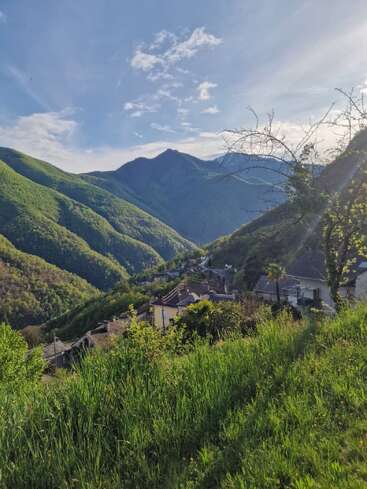 Ein friedliches Bergdorf schmiegt sich in ein üppiges, grünes Tal. Die Häuser stehen verstreut zwischen den Bäumen und das Sonnenlicht fällt durch einen teilweise bewölkten blauen Himmel.