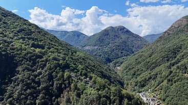 Lush, green mountains dominate the landscape under a bright blue sky with fluffy white clouds. Nestled in the valley below, tiny houses dot the peaceful scenery.