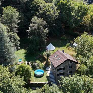 Esta imagen muestra una acogedora casa de piedra con techo rojo, rodeada de frondosos árboles verdes, una pequeña piscina, tiendas de campaña y un tranquilo paisaje boscoso.