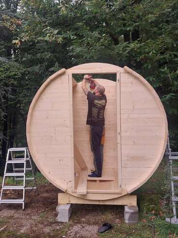 A man is assembling a circular wooden structure, possibly a sauna or tiny cabin, using a power drill. He stands on a platform, surrounded by nature.