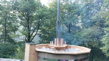A wooden hot tub with bubbling water and a tall metal chimney is set outdoors, surrounded by lush green trees and natural forest scenery, creating tranquility.