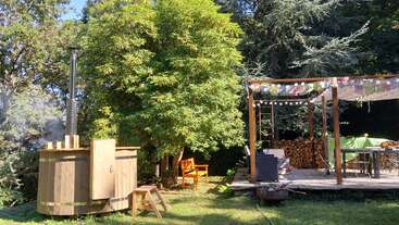 This serene outdoor setting features a wooden hot tub, lush green trees, a shaded wooden patio with seating, fairy lights, stacked firewood, and bright sunshine.
