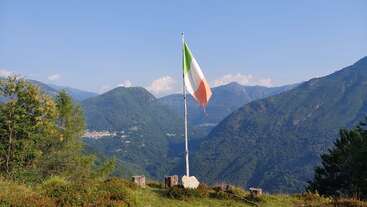 A tall flagpole with the Italian flag stands on a grassy hilltop, surrounded by tree stumps, overlooking lush green mountains under a clear blue sky.