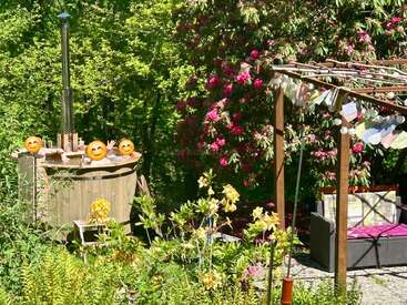In a vibrant garden, three people relax in a wooden hot tub. Nearby, a cozy seating area sits under a pergola adorned with hanging decorations.