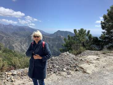 Eine Frau in einem dunklen Mantel und mit Sonnenbrille steht auf einem Bergpfad mit Bäumen. Hinter ihr erstrecken sich sanfte Hügel, ferne Gipfel und ein strahlend blauer Himmel.