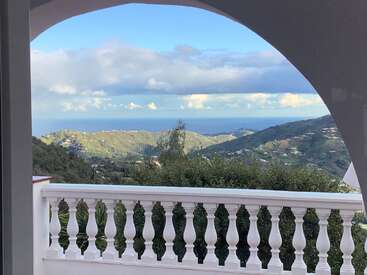 Eine malerische Szene, die durch einen gewölbten Balkon betrachtet wird. Grüne Hügel erstrecken sich bis zum Horizont und treffen auf das blaue Meer unter einem wolkenverhangenen Himmel, der Ruhe ausstrahlt.