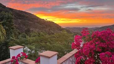 Ein atemberaubender Sonnenuntergang beleuchtet den Himmel orange und gelb über sanften grünen Hügeln, leuchtend rosa Bougainvillea-Blüten und einer charmanten Terrasse mit weißen Wänden. Friedliche Szene.