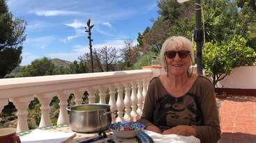 Eine lächelnde Frau mit kurzen weißen Haaren und Sonnenbrille sitzt an einem Tisch im Freien und genießt einen sonnigen Tag, umgeben von Bäumen und schöner Natur.