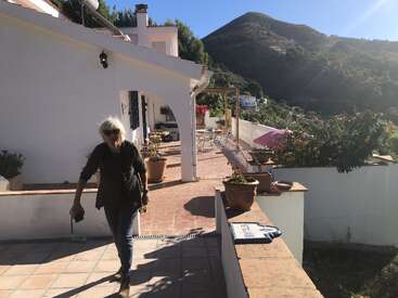Eine Frau spaziert auf einer sonnenbeschienenen Terrasse eines weißen Hauses mit Topfpflanzen, umgeben von grünen Bergen, unter einem klaren blauen Himmel. Friedliche Atmosphäre.