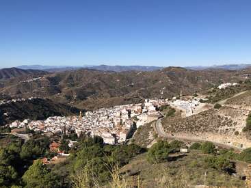 Ein malerisches Dorf mit weißen Häusern schmiegt sich in ein Tal, umgeben von sanften braunen Hügeln und Bergen unter einem klaren blauen Himmel, der von üppigem Grün durchzogen ist.