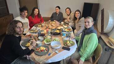 Sieben Freunde sitzen um einen Esstisch herum und essen gemeinsam. Auf dem Tisch stehen verschiedene Speisen, Brot, Gemüse und Getränke. Alle sehen fröhlich und entspannt aus.