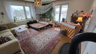 Dieses gemütliche Wohnzimmer verfügt über einen großen Teppich, ein Ecksofa, ein Tagesbett mit Kissen, Pflanzen, ein Klavier, eine warme Beleuchtung und große Fenster, durch die Tageslicht hereinströmt.