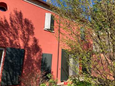 Una casa de un rojo intenso con contraventanas verdes está bañada por la luz del sol. Las sombras de los árboles cercanos se proyectan sobre la pared. El follaje primaveral rodea esta tranquila escena.
