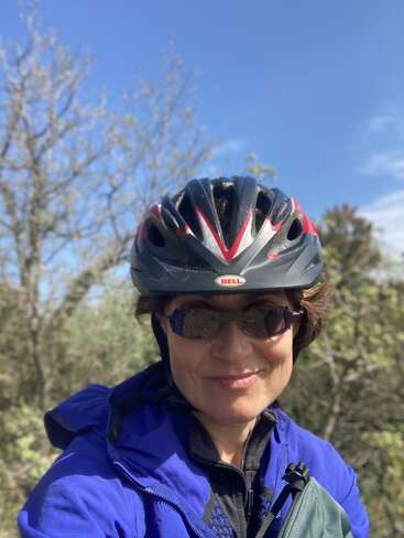 Une femme portant un casque de vélo Bell noir et rouge, des lunettes de soleil et une veste bleue sourit à l'extérieur. L'arrière-plan montre des arbres et un ciel bleu éclatant.