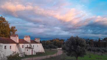 A rustic white farmhouse with red-trimmed doors lies under a dramatic, colorful evening sky. Trees and a winding path surround the peaceful rural landscape scene.